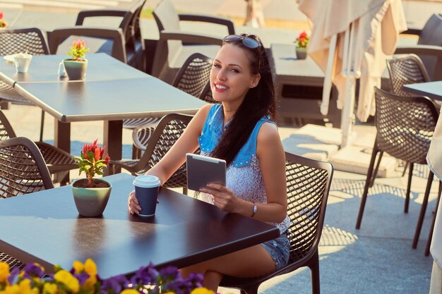 Le portrait d'une belle blogueuse heureuse portant des vêtements à la mode tient une tablette profite de la journée d'été tout en étant assis sur une terrasse dans un café en plein air.