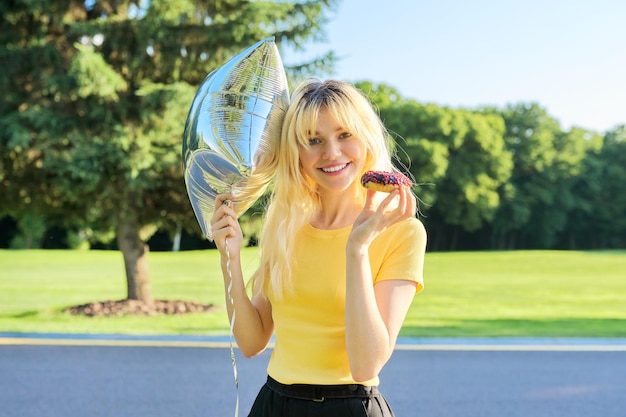 Portrait d'une belle blonde adolescente avec beignet à gâteau et ballon argenté dans le parc