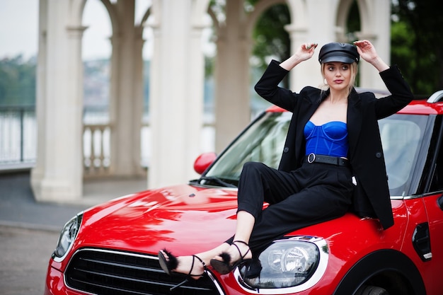 Portrait d'une belle blonde mannequin sexy en casquette et en noir avec un maquillage lumineux assis sur le capot d'une voiture de ville rouge