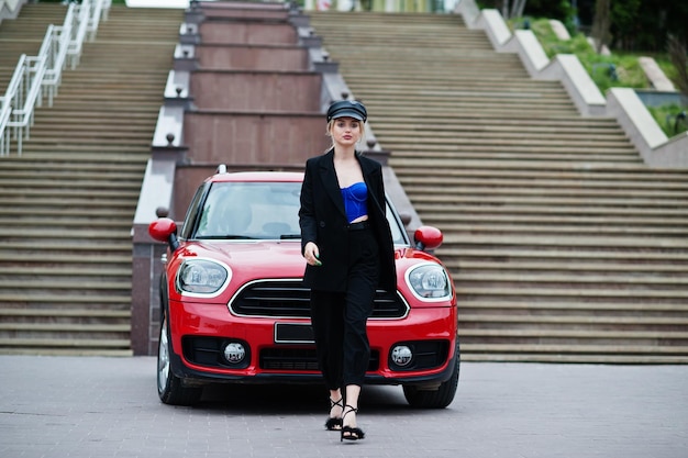 Portrait de la belle blonde sexy mannequin femme en casquette et en corset bleu noir avec un maquillage lumineux près de la voiture de ville rouge