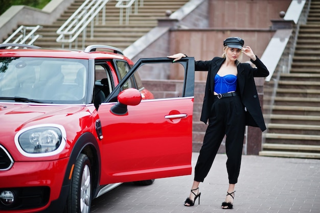 Portrait de la belle blonde sexy mannequin femme en casquette et en corset bleu noir avec un maquillage lumineux près de la voiture de ville rouge