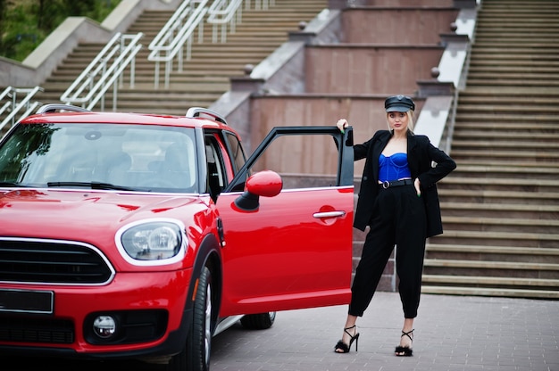 Portrait de la belle blonde sexy mannequin femme en casquette et en tout noir, corset bleu, avec un maquillage lumineux près de la voiture de ville rouge.
