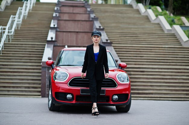 Portrait de la belle blonde sexy mannequin femme en casquette et en tout noir avec un maquillage lumineux près de la voiture de ville rouge.