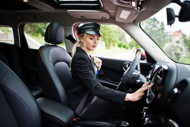 Portrait de la belle blonde sexy mannequin femme en casquette et en tout noir avec un maquillage lumineux s'asseoir et conduire une voiture de ville rouge