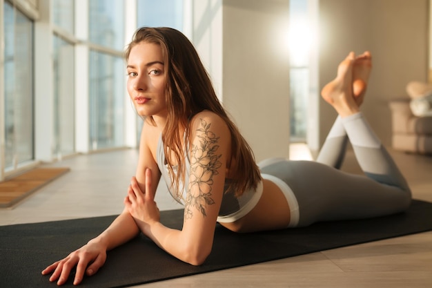 Portrait d'une belle dame en haut sportif et leggings allongé sur un tapis de yoga et regardant pensivement à huis clos à la maison avec de grandes fenêtres en arrière-plan