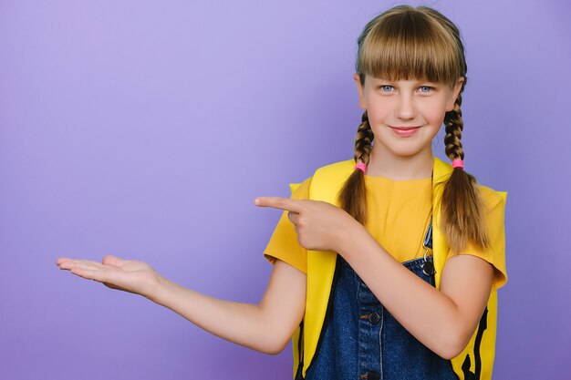 Photo portrait d'une belle écolière intelligente et séduisante portant un sac à dos jaune, posant isolée sur un mur de fond de couleur violette, souriant à la caméra tout en présentant avec la main et en pointant du doigt