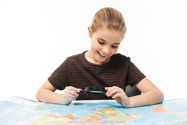 Portrait d'une belle écolière regardant la carte du monde à travers une loupe tout en étudiant la géographie à l'école isolée sur un mur blanc