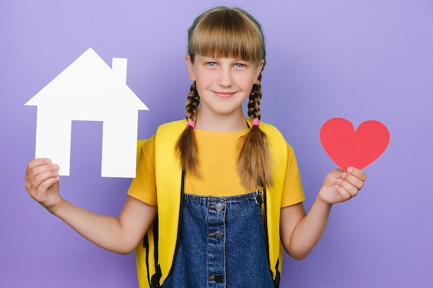 Portrait de belle écolière souriante mignonne tenant un petit coeur rouge et un modèle de maison blanche en papier, heureux de regarder la caméra, porte un sac à dos jaune, isolé sur fond violet pastel en studio