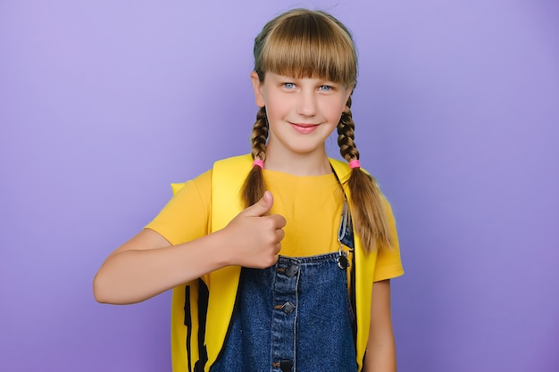 Portrait d'une belle écolière souriante montrant les pouces vers le haut, heureuse de regarder la caméra, porte un sac à dos jaune, posant isolée sur fond de studio violet avec espace de copie. Retour au concept de l'école