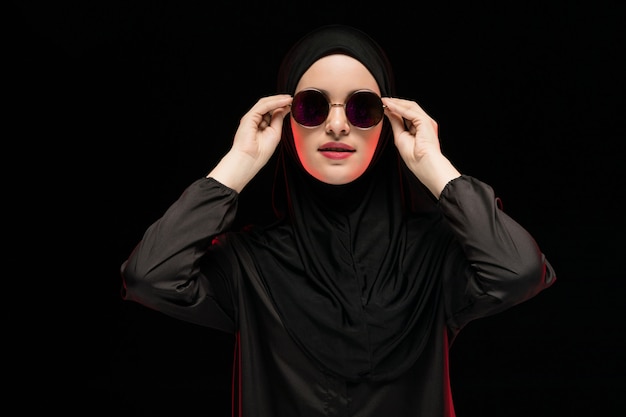 Portrait de la belle élégante jeune femme musulmane portant le hijab noir et des lunettes de soleil comme concept de mode orientale moderne posant sur fond noir