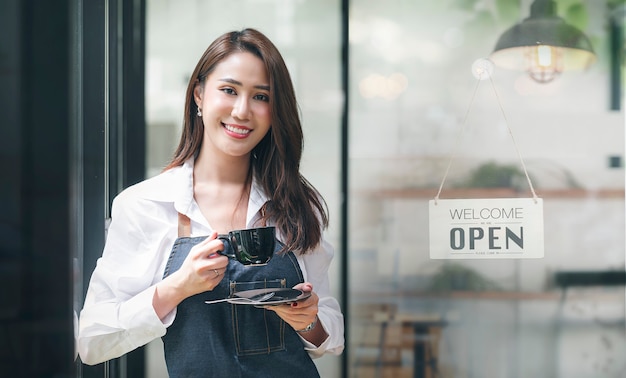 Portrait d'une belle entrepreneure asiatique tenant une tasse debout à la porte de sa boutique avec un panneau ouvert.