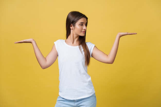 Portrait d'une belle étudiante, pigiste insensée, bonne épouse positive, fille sympathique regardant la caméra isolée sur fond jaune gesticulant des espaces vides des deux côtés avec des paumes.