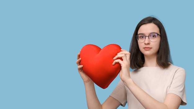 Portrait d'une belle étudiante souriante avec des lunettes tenant un oreiller en forme de coeur rouge vif