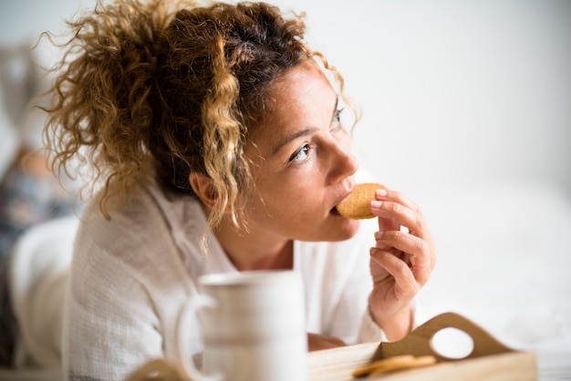 Portrait de belle femme adulte mangeant le biscuit dans le petit déjeuner de matin dans la chambre à coucher
