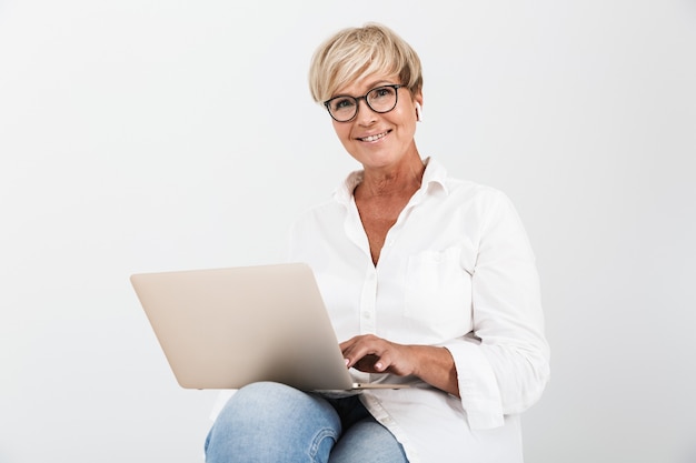 Portrait d'une belle femme adulte portant des lunettes et des écouteurs assis avec un ordinateur portable isolé sur un mur blanc en studio