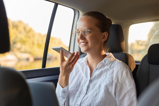 Portrait d'une belle femme d'affaires caucasienne positive et optimiste souriante utilisant un ordinateur portable et un téléphone portable assis sur la banquette arrière d'une voiture enregistrant un message vocal