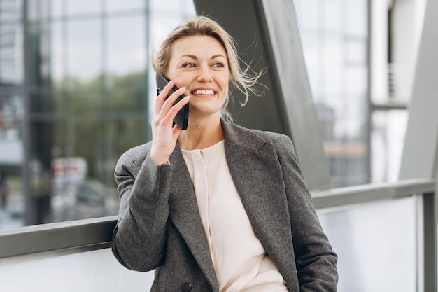 Portrait d'une belle femme d'affaires mature en costume et veste grise souriant et parlant au téléphone sur le fond urbain moderne