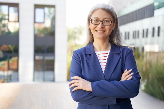 Portrait d'une belle femme d'affaires mature et heureuse en lunettes et vêtements classiques gardant les bras