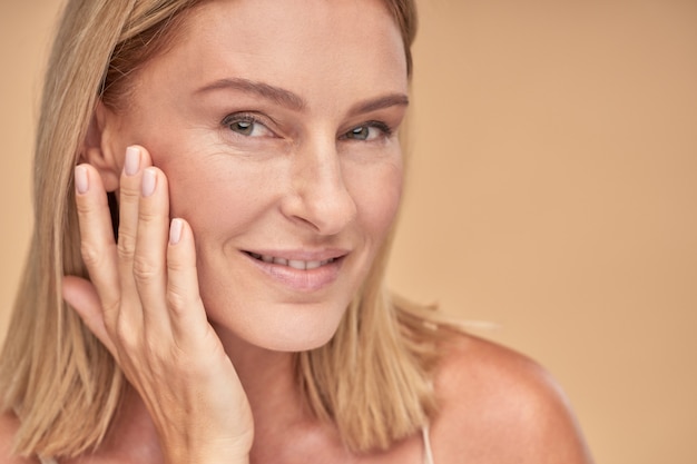 Photo portrait d'une belle femme d'âge moyen touchant sa peau propre et souriant à la caméra tout en