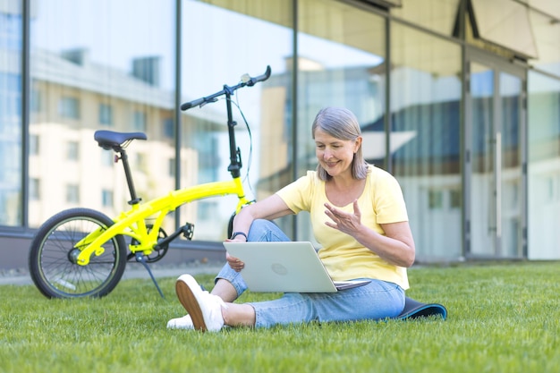 Portrait d'une belle femme âgée assise sur le campus sur l'herbe avec un ordinateur portable et un vélo Parler lors d'un appel vidéo sur un ordinateur portable en agitant les bras en souriant