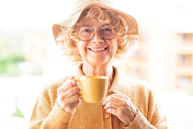 Portrait d'une belle femme âgée bouclée heureuse en jaune avec chapeau et lunettes de soleil tenant une tasse de café en regardant la caméra