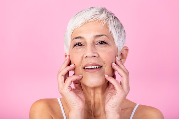 Portrait d'une belle femme âgée touchant sa peau parfaite et regardant la caméra Visage en gros plan d'une femme mature avec des rides massant le visage isolé sur un fond rose Concept du processus de vieillissement