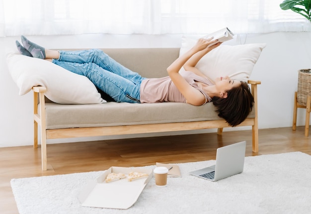 Portrait d'une belle femme asiatique lisant un livre et allongée sur le canapéfaites une pause en mangeant de la pizza et travaillez à la maison pendant l'auto-isolement de la crise de l'épidémie de coronavirusNouveau concept de mode de vie normal