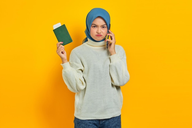 Portrait d'une belle femme asiatique pensive en chandail blanc parlant sur un téléphone portable et détenant un passeport isolé sur fond jaune Voyage d'été