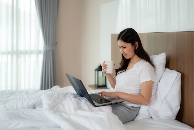 Portrait de la belle femme asiatique se détendre à l'aide d'un ordinateur portable et boire du café dans la chambre. Sourire heureuse fille asiatique shopping en ligne à la maison Education e-commerce freelance technologie connectée asie personnes concept