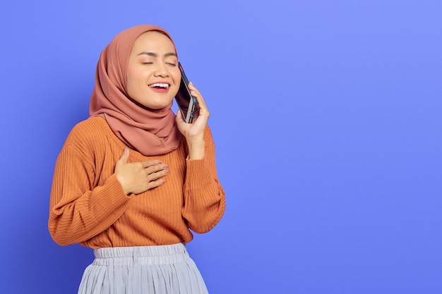 Portrait d'une belle femme asiatique souriante en chandail marron et hijab parlant au téléphone portable avec la main sur la poitrine isolée sur fond violet Concept religieux de l'islam des gens