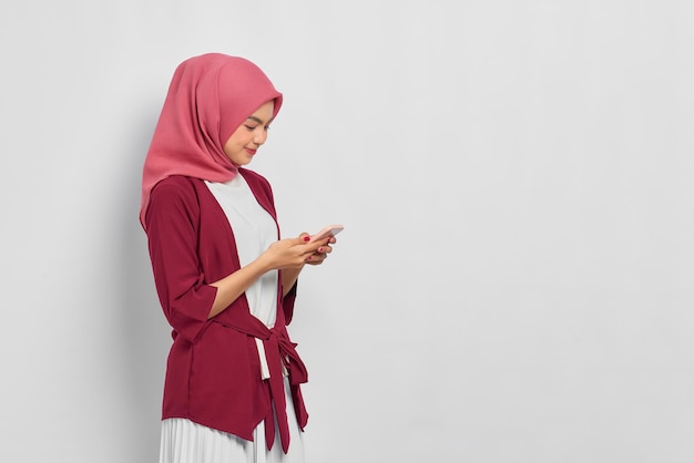 Portrait D'une Belle Femme Asiatique Souriante En Chemise Décontractée Et Hijab à L'aide D'un Téléphone Portable, Tapant Des Messages Sms Avec Espace De Copie Isolé Sur Fond Blanc. Concept De Mode De Vie Religieux De Personnes