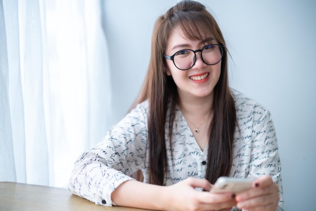 Photo portrait d'une belle femme asiatique souriante et heureuse relaxante écrivant un message sur son smartphone assise sur le canapé sur un canapé confortable à la maison dans le salon