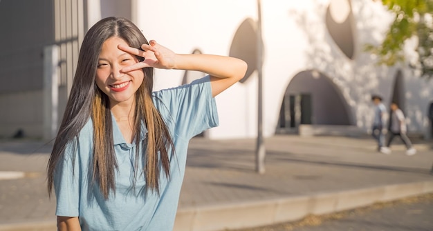 Portrait d'une belle femme asiatique souriante et regardant les doigts de la victoire de la caméra à l'extérieur