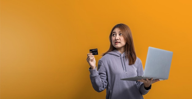 Portrait d'une belle femme asiatique tenant une carte de crédit et un ordinateur portable sur le concept de portrait de fond jaune isolé utilisé pour la publicité et la signalisation isolée sur l'espace de copie de fond bleu