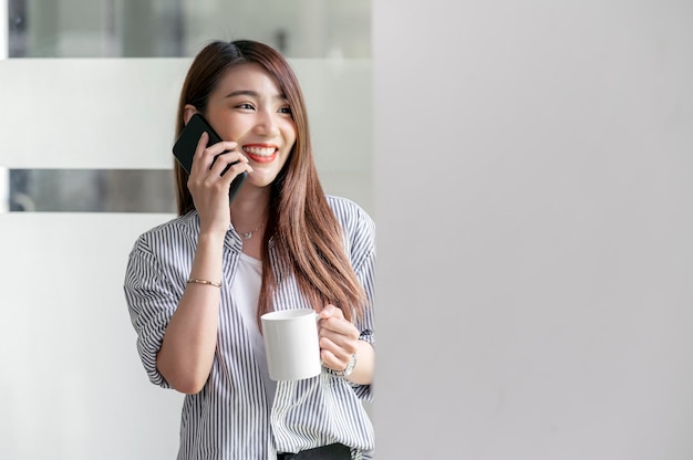 Portrait d'une belle femme asiatique tenant une tasse et utilisant un smartphone avec bonheur en se tenant debout au bureau.