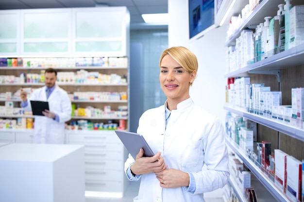 Portrait de la belle femme blonde pharmacien debout dans la pharmacie ou la pharmacie par l'étagère avec des médicaments et tenant la tablette.
