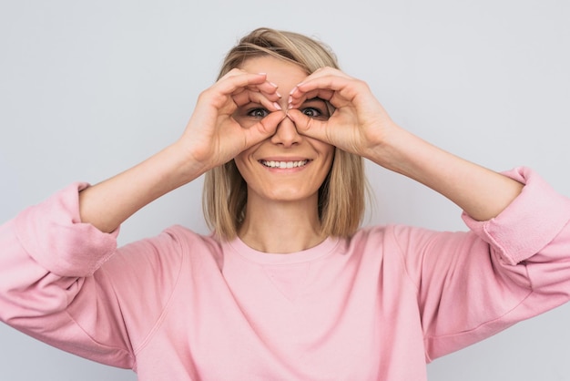 Portrait d'une belle femme blonde portant un pull décontracté rose tenant ses mains à ses yeux comme si elle regardait à travers des jumelles ou des lunettes Surpris femme heureuse sur fond de mur blanc de studio