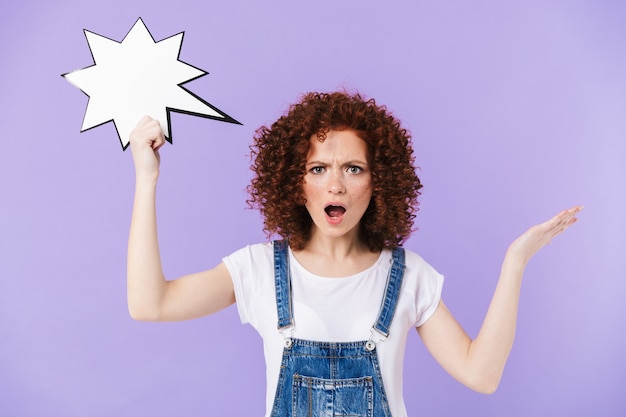 Portrait d'une belle femme bouclée rousse des années 20 souriant tout en tenant une bulle de pensée vierge isolée sur un mur violet