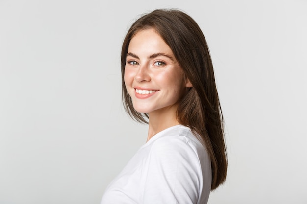 Portrait de belle femme brune confiante tournant le visage à la caméra avec un regard rêveur, souriant sur blanc.