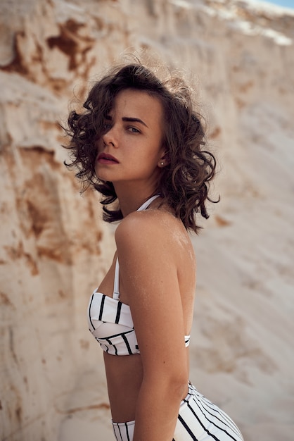 Portrait de la belle femme caucasienne bronzée modèle aux cheveux longs noirs en maillot de bain rayé allongé sur la plage d'été avec du sable blanc. Vue de dessus