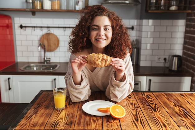 Portrait d'une belle femme caucasienne mangeant un croissant et buvant du jus d'orange à table à l'intérieur de la cuisine tout en prenant son petit déjeuner à la maison