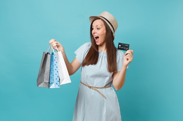 Portrait d'une belle femme caucasienne en robe d'été, chapeau de paille tenant des sacs de paquets avec des achats après le shopping, carte de crédit bancaire isolée sur fond bleu pastel. Copiez l'espace pour la publicité.