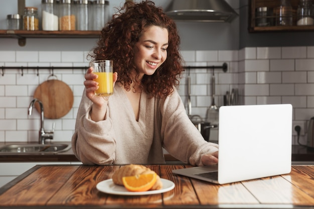 Portrait d'une belle femme caucasienne utilisant un ordinateur portable à table à l'intérieur de la cuisine tout en prenant son petit-déjeuner à la maison