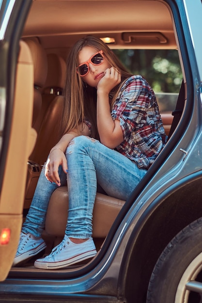 Portrait d'une belle femme en chemise polaire et jeans, assise dans la voiture sur le siège arrière avec une porte ouverte.