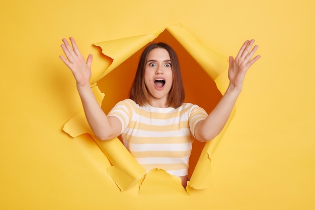 Portrait d'une belle femme excitée portant un t-shirt rayé montrant une grande taille avec les mains regardant la caméra avec une expression étonnée regardant à travers la percée du fond jaune