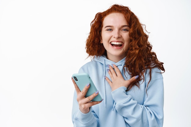 Portrait d'une belle femme heureuse aux cheveux roux bouclés, riant après avoir lu un message sur un smartphone, tenant un téléphone portable et souriant sur blanc