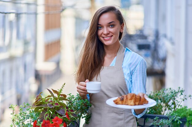 Portrait d'une belle femme heureuse mignonne joyeuse souriante chef pâtissier avec une assiette de croissants bruns frais cuits au four et tasse de café