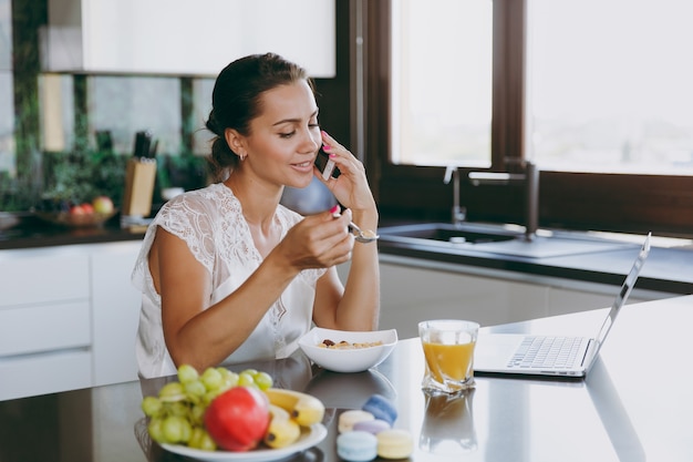 Le portrait d'une belle femme heureuse parlant sur un téléphone portable pendant le petit-déjeuner avec un ordinateur portable sur la table