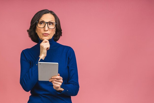 Portrait d'une belle femme mature âgée à l'aide d'un ordinateur tablette, isolée sur fond rose. Copiez l'espace pour le texte.