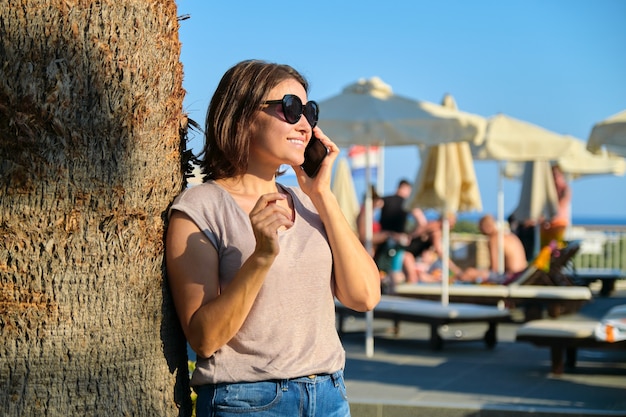 Portrait d'une belle femme mature souriante se relaxant dans un hôtel de villégiature, femme à lunettes de soleil debout près d'un palmier à l'aide d'un smartphone, espace pour copie Vacances, été, tourisme, station balnéaire, personnes d'âge moyen